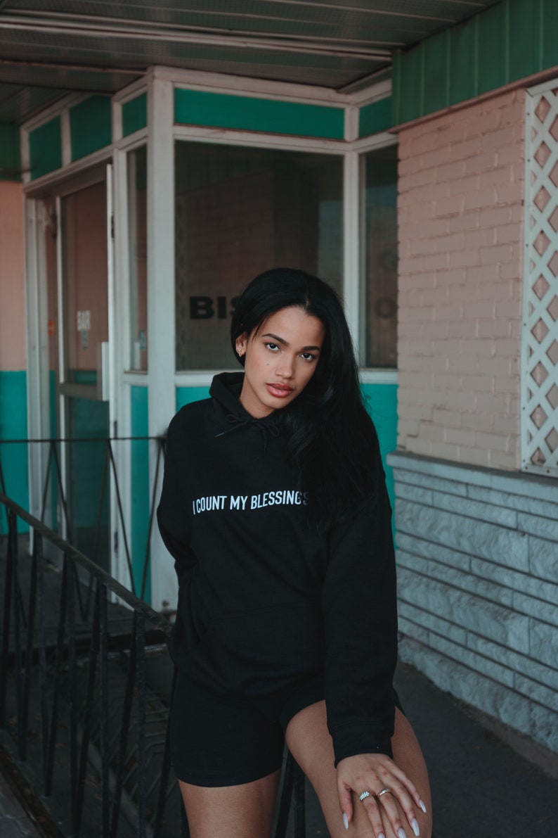 K&ouml;nnte beinhalten: Schwarzer Kapuzenpullover mit dem wei&szlig;en Text "I COUNT MY BLESSINGS". Das Model tr&auml;gt schwarze Shorts. Der Hintergrund zeigt ein Geb&auml;ude mit einer t&uuml;rkisfarbenen und rosa Fassade.