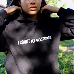 K&ouml;nnte beinhalten: Schwarzer Kapuzenpullover mit dem wei&szlig;en Text "I COUNT MY BLESSINGS". Der Hoodie hat eine Vordertasche und eine Kapuze mit Kordelzug. Die Person tr&auml;gt den Hoodie im Freien.