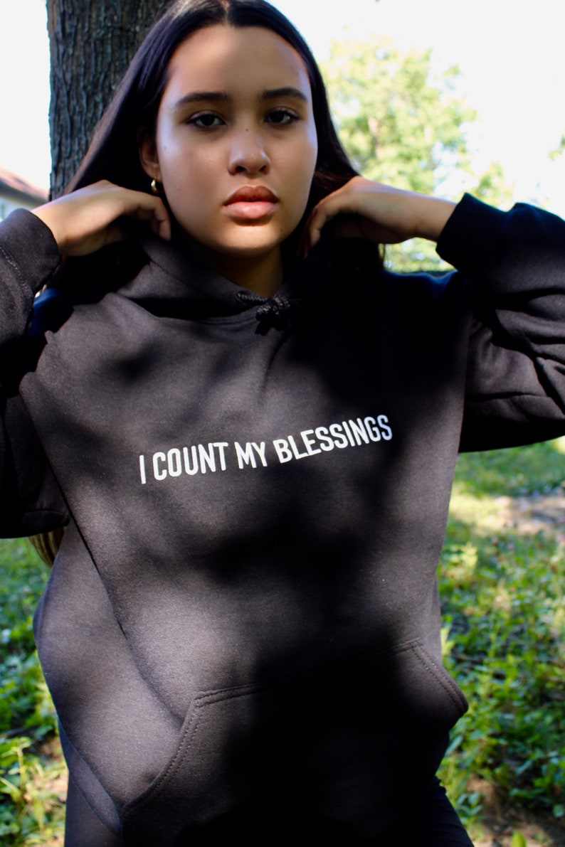 K&ouml;nnte beinhalten: Schwarzer Kapuzenpullover mit dem wei&szlig;en Text "I COUNT MY BLESSINGS". Der Hoodie hat eine Vordertasche und eine Kapuze mit Kordelzug. Die Person tr&auml;gt den Hoodie im Freien.