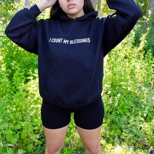 K&ouml;nnte beinhalten: Schwarzer Kapuzenpullover mit dem wei&szlig;en Text "I COUNT MY BLESSINGS". Der Hoodie hat eine Vordertasche und eine Kapuze mit Kordelzug. Die Person tr&auml;gt schwarze Shorts und wei&szlig;e Turnschuhe. Der Hintergrund ist eine nat&uuml;rliche Umgebung im Freien.