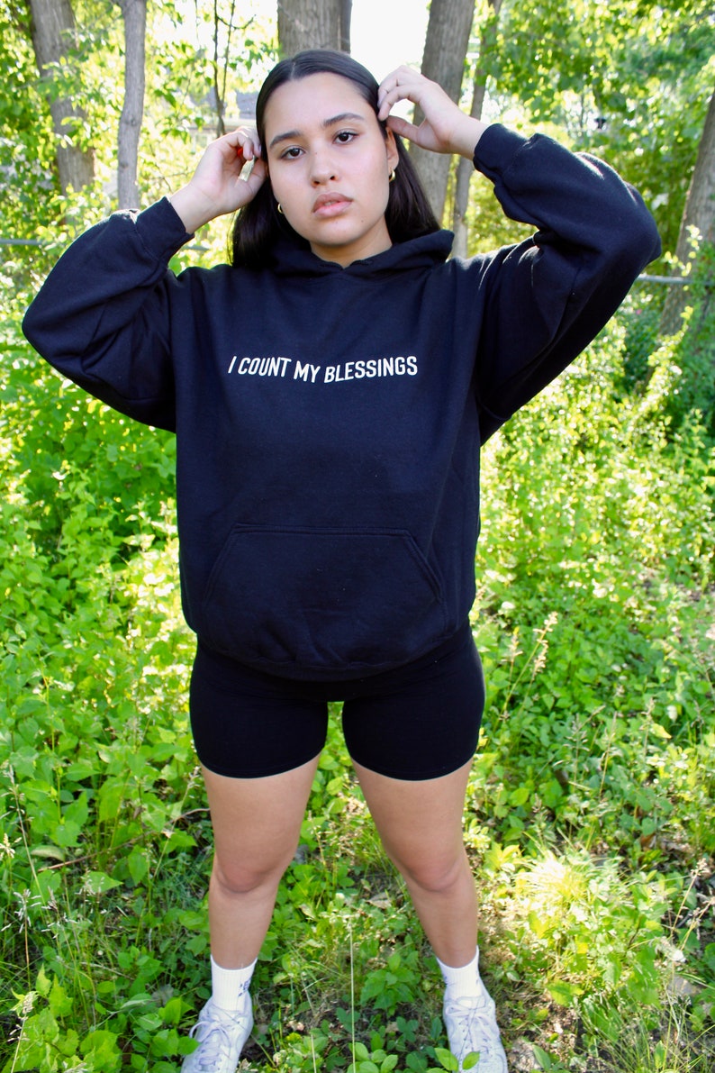 K&ouml;nnte beinhalten: Schwarzer Kapuzenpullover mit dem wei&szlig;en Text "I COUNT MY BLESSINGS". Der Hoodie hat eine Vordertasche und eine Kapuze mit Kordelzug. Die Person tr&auml;gt schwarze Shorts und wei&szlig;e Turnschuhe. Der Hintergrund ist eine nat&uuml;rliche Umgebung im Freien.