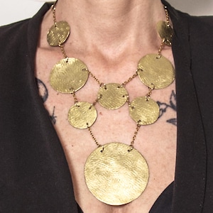 May include: A gold-toned necklace featuring multiple circular pendants of varying sizes. The pendants are connected by delicate chains, creating a layered effect. The necklace is worn against a dark background, highlighting its metallic sheen.