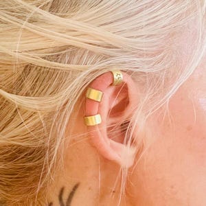 Può includere: Tre ear cuff color oro indossati sulla cartilagine superiore. Il cerchietto superiore presenta un piccolo motivo a stella inciso, mentre i due posizionati sotto sono lisci, semplici e di forma rettangolare, avvolgendo il bordo esterno dell'orecchio.