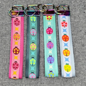 May include: Four key fobs with ladybug designs. Each fob features a different color scheme: pink, teal, light blue, and gray. The ladybugs are in various colors, including red, yellow, and purple. Each fob has a metal clasp.