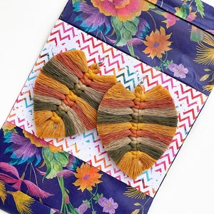 Puede incluir: Un par de pendientes de macramé hechos a mano, en forma de hoja, en tonos amarillo mostaza, naranja y verde oliva. Se encuentran sobre un fondo con diseños florales y zigzags.