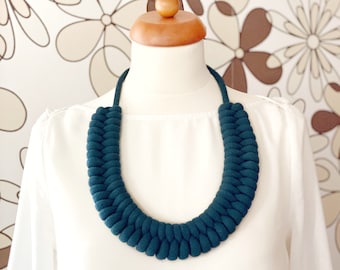Op de afbeelding: Een donkerblauwe stoffen ketting met een gevlochten ontwerp. De ketting wordt getoond op een wit mannequin.