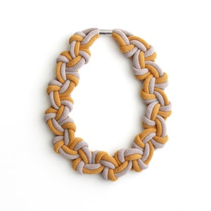 Puede incluir: Un collar anudado hecho de dos tonos de hilo, uno amarillo mostaza y el otro gris claro. El collar est&aacute; atado en un bucle continuo con un cierre met&aacute;lico.
