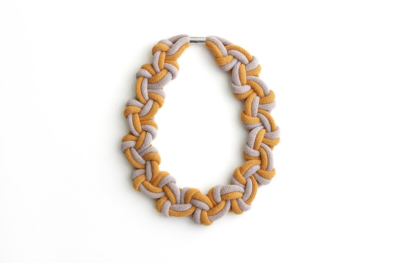 Puede incluir: Un collar anudado hecho de dos tonos de hilo, uno amarillo mostaza y el otro gris claro. El collar est&aacute; atado en un bucle continuo con un cierre met&aacute;lico.
