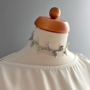 Può includere: Una delicata collana di foglie all'uncinetto in tonalità di verde e grigio, esposta su un manichino bianco con una testa di legno.