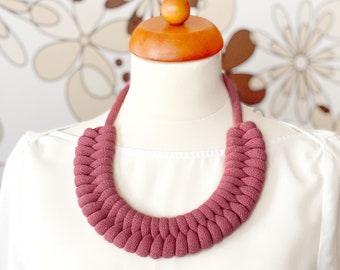 Op de afbeelding: Een dikke, gevlochten ketting gemaakt van een dieproze stof. De ketting wordt getoond op een wit mannequin met een houten hoofd.