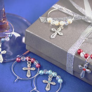 May include: Wine glass charms with silver crosses and colorful beads. The charms are attached to silver hoops and are displayed on a blue surface next to a silver gift box with a ribbon.