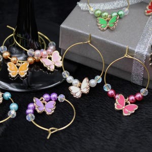 May include: A collection of colorful butterfly-themed wine charms. Each charm features a gold-tone hoop with a butterfly pendant and decorative beads in various colors, including pink, purple, and yellow. The charms are displayed on a black surface.