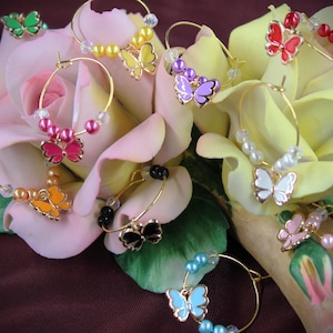 Puede incluir: Pendientes de aro dorados con colgantes de mariposa en varios colores, como rosa, amarillo, morado y azul. Los pendientes están adornados con cuentas y se exhiben sobre un lecho de flores rosas y amarillas. Un accesorio delicado.