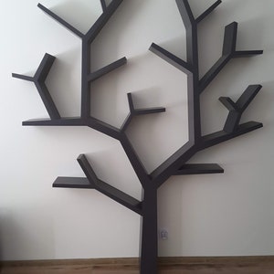May include: A dark gray tree-shaped wall shelf with multiple branches and shelves. The shelves are designed to resemble the branches of a tree, creating a unique and decorative storage solution. The shelf is mounted on a white wall above a wooden floor.