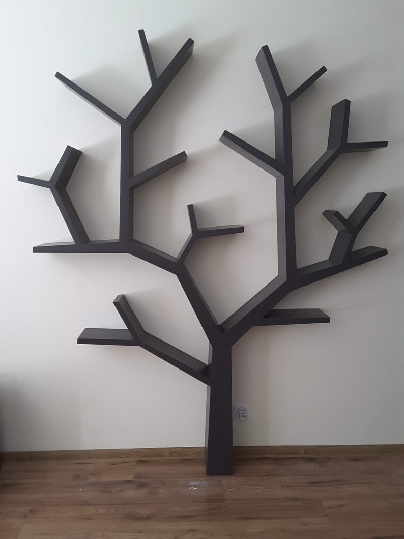 May include: A dark gray tree-shaped wall shelf with multiple branches and shelves. The shelves are designed to resemble the branches of a tree, creating a unique and decorative storage solution. The shelf is mounted on a white wall above a wooden floor.