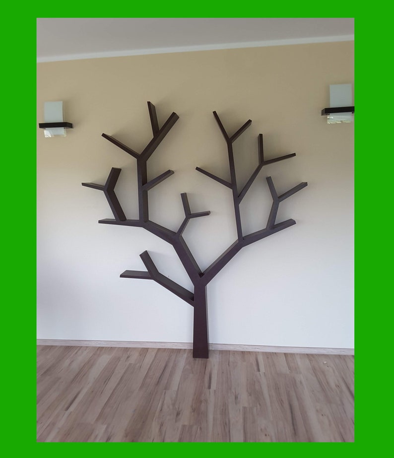 May include: A dark brown tree-shaped bookshelf mounted on a beige wall. The bookshelf has multiple branches with small shelves, and is positioned above a wooden floor. Two wall sconces are on either side of the bookshelf.
