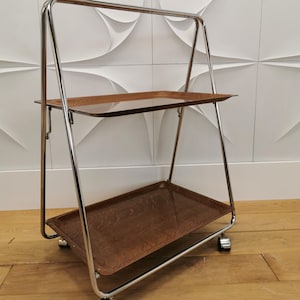 Könnte beinhalten: Ein verchromter Servierwagen mit zwei Holztabletts. Der Wagen hat einen rechteckigen Rahmen und steht auf vier Rädern. Die Tabletts sind dunkelbraun. Der Wagen steht vor einer strukturierten weißen Wand.