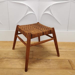 Könnte beinhalten: Ein kleiner Holzschemel mit geflochtener Sitzfläche. Der Hocker hat vier angewinkelte Beine und eine rechteckige Sitzfläche aus geflochtenem Material. Das Holz hat einen warmen, braunen Farbton. Ein funktionelles Möbelstück.