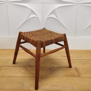 Könnte beinhalten: Kleiner, quadratischer Holzschemel mit geflochtener Sitzfläche. Der Hocker hat vier angewinkelte Beine und eine dunkelbraune Oberfläche. Der Sitz besteht aus geflochtenen Naturfasern. Es ist ein Vintage- oder antikes Stück.