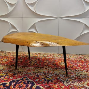 Könnte beinhalten: Ein moderner Couchtisch aus der Mitte des Jahrhunderts mit einer Tischplatte aus Naturholz und schwarzen Metallbeinen. Die organische Form und die warmen Holztöne stehen im Kontrast zur weißen Wand. Der Tisch ist etwa 122 cm lang.