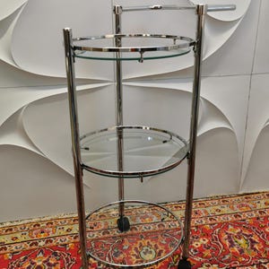 Könnte beinhalten: Ein dreistöckiger Servierwagen aus Chrom und Glas mit Rollen. Der Wagen hat eine runde Oberseite und zwei untere Ablagen.