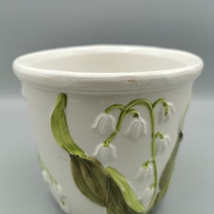 Könnte beinhalten: Weißer Keramik-Blumentopf mit geprägtem Maiglöckchen-Design. Der Topf hat grüne Blätter und weiße, glockenförmige Blüten. Der Topf hat eine abgerundete Form mit einem leicht ausgestellten Rand.