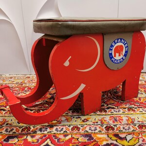 Könnte beinhalten: Ein roter Elefantenhocker aus Holz mit einem grauen gepolsterten Sitz. Der Elefant hat eine weiße Umrandung und die Worte "Elefanten Marke" sind an der Seite aufgedruckt.