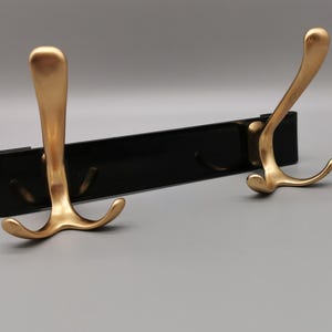 Könnte beinhalten: Ein schwarzer Wandgarderobenständer mit drei goldfarbenen Haken. Die Haken haben ein geschwungenes Design und sind an einer rechteckigen schwarzen Basis befestigt. Der Ständer ist zum Aufhängen von Mänteln, Hüten oder anderen Gegenständen konzipiert.