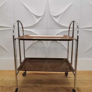Könnte beinhalten: Ein brauner Servierwagen mit zwei rechteckigen Tabletts. Der Wagen hat ein verchromtes Gestell und kleine Räder. Die Tabletts haben eine Holzmaserung. Der Wagen steht vor einer weißen strukturierten Wand.