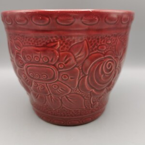 Könnte beinhalten: Ein roter Keramik-Blumentopf mit geprägten floralen Mustern. Der Topf weist ein sich wiederholendes Design aus Blumen und Blättern mit einer strukturierten Oberfläche auf. Der Rand hat eine dekorative Bordüre aus kleinen, abgerundeten Formen. Dieser dekorative Topf ist für den Innen- oder Außenbereich geeignet.