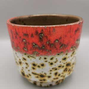 Könnte beinhalten: Eine Keramiktasse mit einem leuchtend roten Oberteil und einem gesprenkelten weißen und braunen unteren Bereich. Der Innenrand ist dunkelbraun. Die Tasse hat eine abgerundete Form und einen leicht ausgestellten Rand. Das Design ist eine Mischung aus Texturen und Farben.