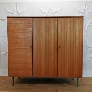 Könnte beinhalten: Ein rechteckiger Holzschrank mit drei vertikalen Türen in natürlicher Maserung. Das Möbelstück steht auf vier dünnen, geraden Metallbeinen. Jede Tür ist mit einem kleinen, quadratischen Metallgriff ausgestattet, der mittig am Rand sitzt.