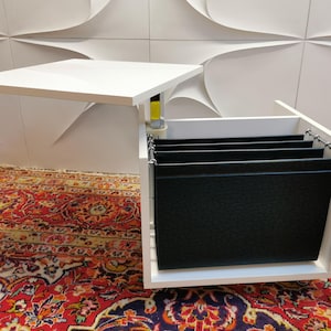 Könnte beinhalten: Ein weißer Schreibtisch mit einer hochklappbaren Oberseite, die ein Fach mit schwarzen Hängeregistern freigibt.