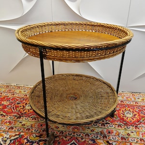 Könnte beinhalten: Ein zweistöckiger Servierwagen mit einer runden Ober- und Unterseite aus Weide. Die obere Ebene hat eine Holzoberfläche und die untere Ebene ist offen. Der Wagen ist aus schwarzem Metall und hat Räder.