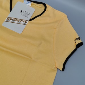 Könnte beinhalten: Ein gelbes T-Shirt mit kurzen Ärmeln und schwarzem Besatz an Kragen und Ärmeln. Das Shirt hat ein Etikett mit dem Wort "SPORTING" und anderem Text. Auf dem Ärmel steht das Wort "SPORT".