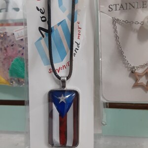May include: A black leather cord necklace with a silver pendant featuring the Puerto Rican flag. The pendant is rectangular and has a clear glass front. The flag is red, white, and blue with a white star in the blue triangle.