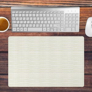 Op de afbeelding: Bovenaanzicht van een werkplek met een zilverkleurig toetsenbord, een witte muis en een kopje koffie op een donkerhouten oppervlak. Een crèmekleurige muismat met horizontale lijnen staat in het midden.