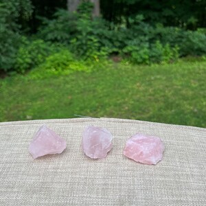 May include: Three pink rose quartz crystals on a beige textured surface. The crystals are rough and have a natural, unpolished look.