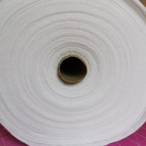 May include: A roll of white, fluffy fabric, tightly wound. The fabric appears to be a soft, non-woven material, possibly batting or interfacing. The core is a brown cardboard tube.
