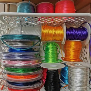 May include: A white plastic basket filled with spools of colorful satin cord and stacked discs of various colored cord. The colors include teal, pink, red, yellow, purple, orange, green, black, blue, and silver. The cords are used for crafting and jewelry making.