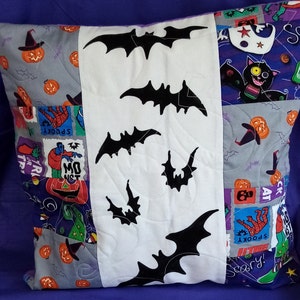 May include: A white pillow with a patchwork design featuring various Halloween-themed images, including pumpkins, bats, and witches. The pillow has a white panel in the centre with black bat silhouettes.