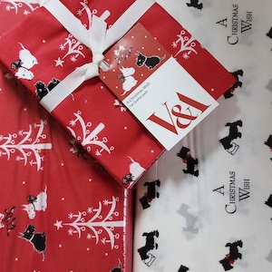 May include: A wrapped gift with a white ribbon and tag rests on red wrapping paper decorated with white Christmas trees and dog illustrations. The tag reads "A Christmas Wish" and features the V&A logo. Additional wrapping paper with black dog illustrations and the text "A Christmas Wish" is visible.