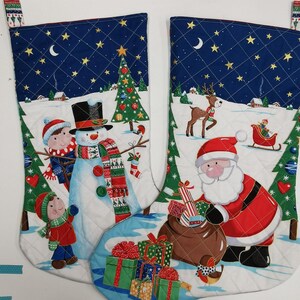 May include: Two Christmas stockings with quilted white fabric and a blue top. One stocking features a snowman, children, and a Christmas tree. The other stocking shows Santa Claus, a reindeer, and a sleigh.