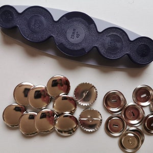 May include: A set of silver-coloured metal button parts for making custom buttons. The set includes a variety of sizes, including 19mm, 25mm, and 29mm. The button parts are arranged on a white surface, along with a blue plastic tool for attaching the buttons.