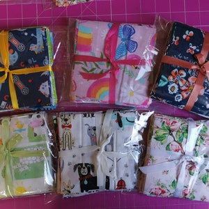 May include: Fabric bundles in clear packaging, tied with ribbons. Patterns include dinosaurs, dogs, flowers, and rainbows. Ribbons are yellow, pink, and peach. The bundles are arranged on a pink surface. Each bundle contains several pieces of fabric.