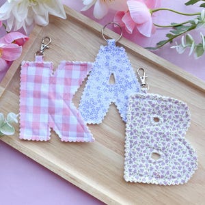 Peut inclure: Trois porte-clés en tissu en forme de lettres : un "K" à carreaux roses, un "A" floral lavande et un "B" floral lavande. Chaque lettre a un fermoir argenté et un anneau métallique. Les porte-clés sont sur un plateau en bois avec des fleurs roses et blanches.