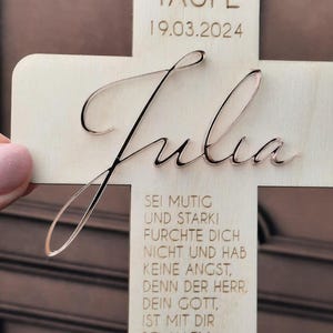 May include: A wooden cross with the name "Julia" engraved on it. The cross is decorated with a small fish symbol and a message in German that reads "Sei mutig und stark! Furchte dich nicht und hab keine Angst, denn der Herr, dein Gott, ist mit dir bei allem, was du unternimmst." The date "19.03.2024" is also engraved on the cross.