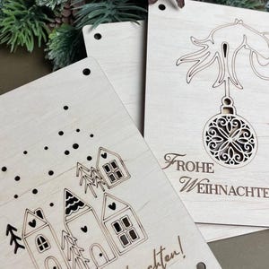 May include: Two wooden Christmas card designs. The first card features a simple design of three houses with snow falling. The second card features a Christmas tree with a hanging ornament and the text "Frohe Weihnachten!" The text at the bottom of the cards reads "SVG - DXF - PNG - PDF instant file download".
