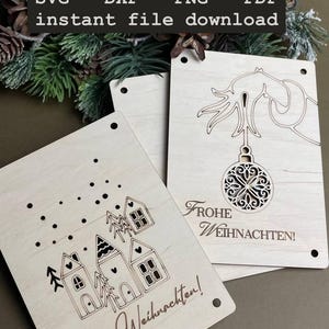 May include: Two wooden Christmas cards with laser cut designs. One card features a Christmas tree with an ornament and the text "Frohe Weihnachten!" The other card features a row of houses with the text "Frohe Weihnachten!"
