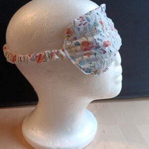 May include: A floral sleep mask with a white base, adorned with red, orange, and blue flower patterns. The mask includes an elasticated band. This sleep mask is designed to block out light for improved sleep.